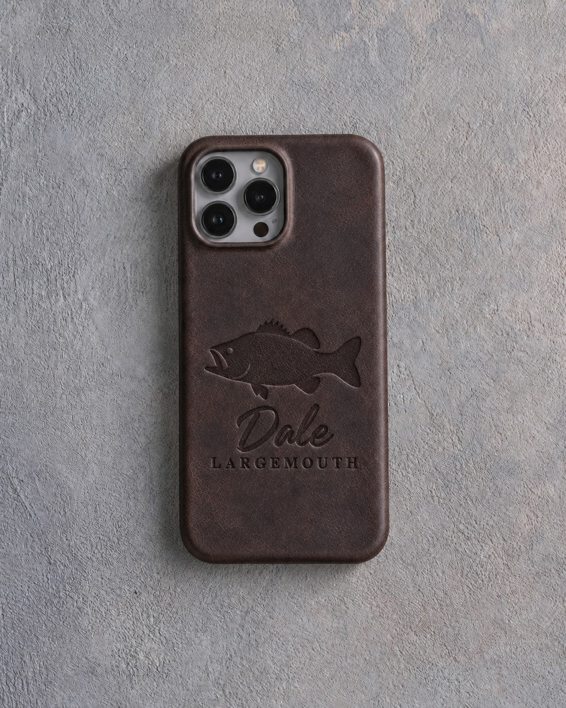 Back of a dark espresso leather iPhone case debossed with a largemouth bass above Dale and LARGEMOUTH below the camera, on gray plaster