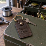 Dark espresso Dale LARGEMOUTH keychain in full-grain leather on the lid of an olive-green steel tackle box in a garage, lures in the trays behind