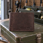Dark espresso Dale LARGEMOUTH bifold wallet in full-grain leather on the lid of an olive-green steel tackle box in a garage, lures in the trays behind
