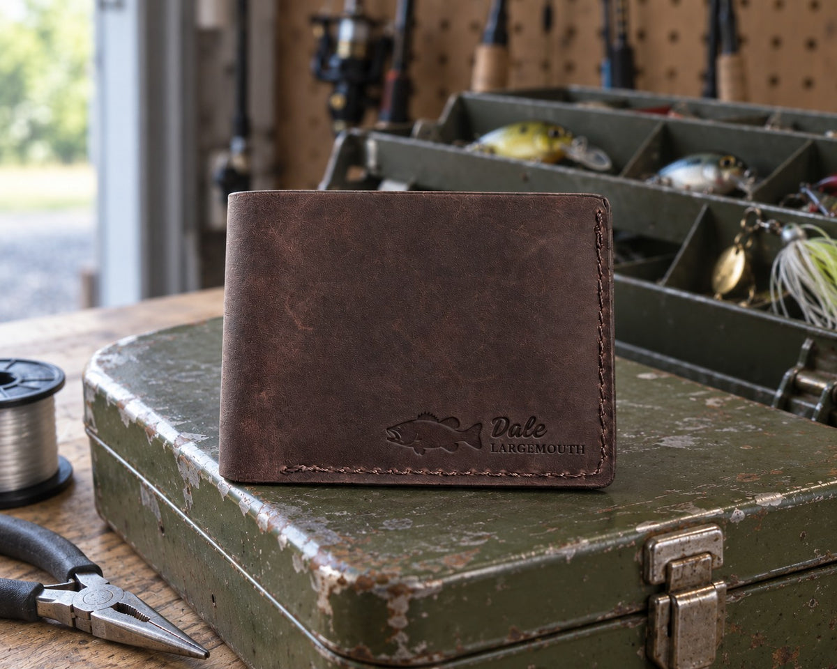 Dark espresso Dale LARGEMOUTH bifold wallet in full-grain leather on the lid of an olive-green steel tackle box in a garage, lures in the trays behind