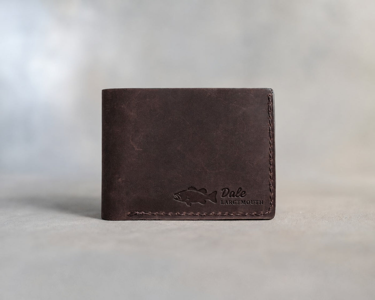 Closed dark espresso leather bifold wallet with a largemouth bass beside Dale and LARGEMOUTH pressed in the lower right corner, on gray plaster
