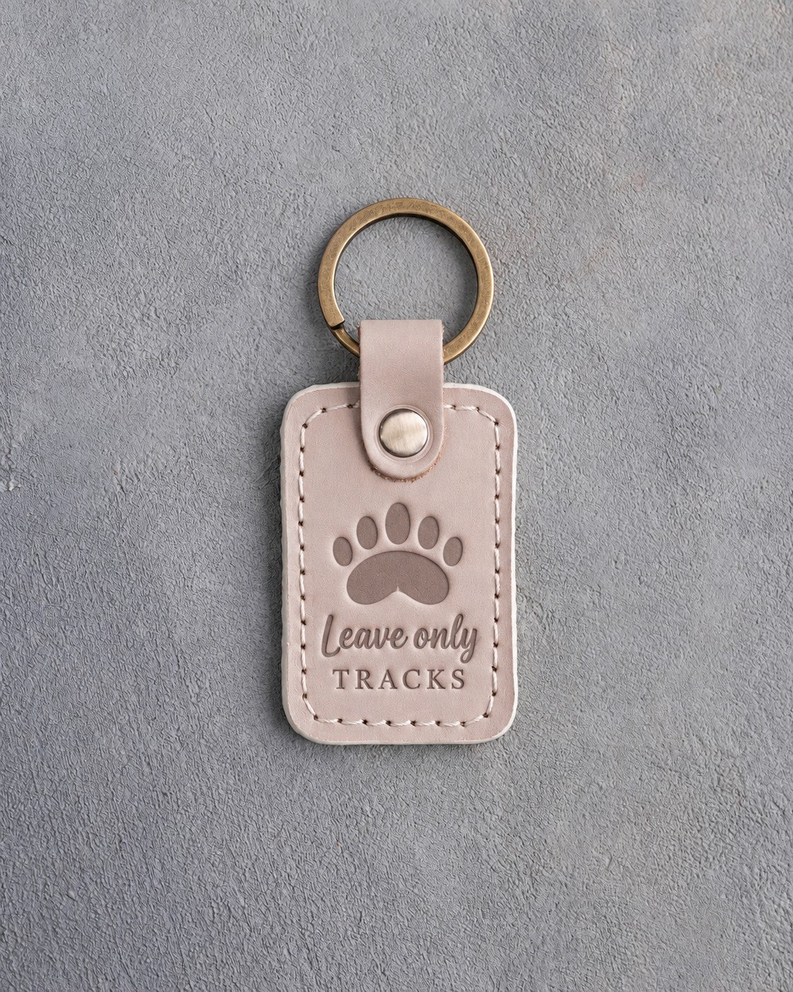 Gray sand leather keychain debossed with a black bear paw track above the words Leave only TRACKS, on gray plaster