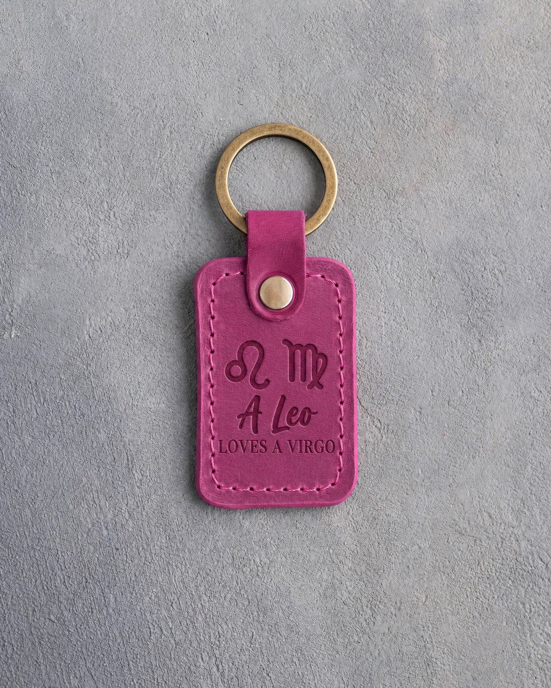 Fuchsia pink leather keychain debossed with Leo and Virgo zodiac glyphs above A Leo and LOVES A VIRGO, on gray concrete