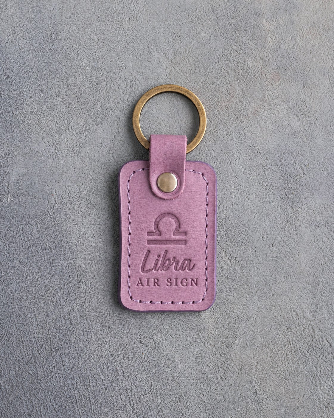 Lilac leather keychain debossed with the Libra zodiac glyph above Libra and AIR SIGN, on gray concrete