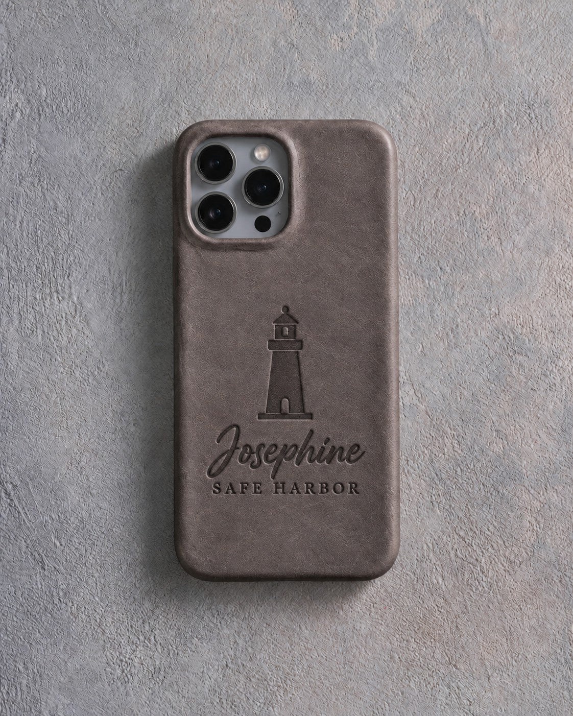 Back of a country gray leather iPhone case debossed with a lighthouse above Josephine and SAFE HARBOR below the camera, on gray plaster