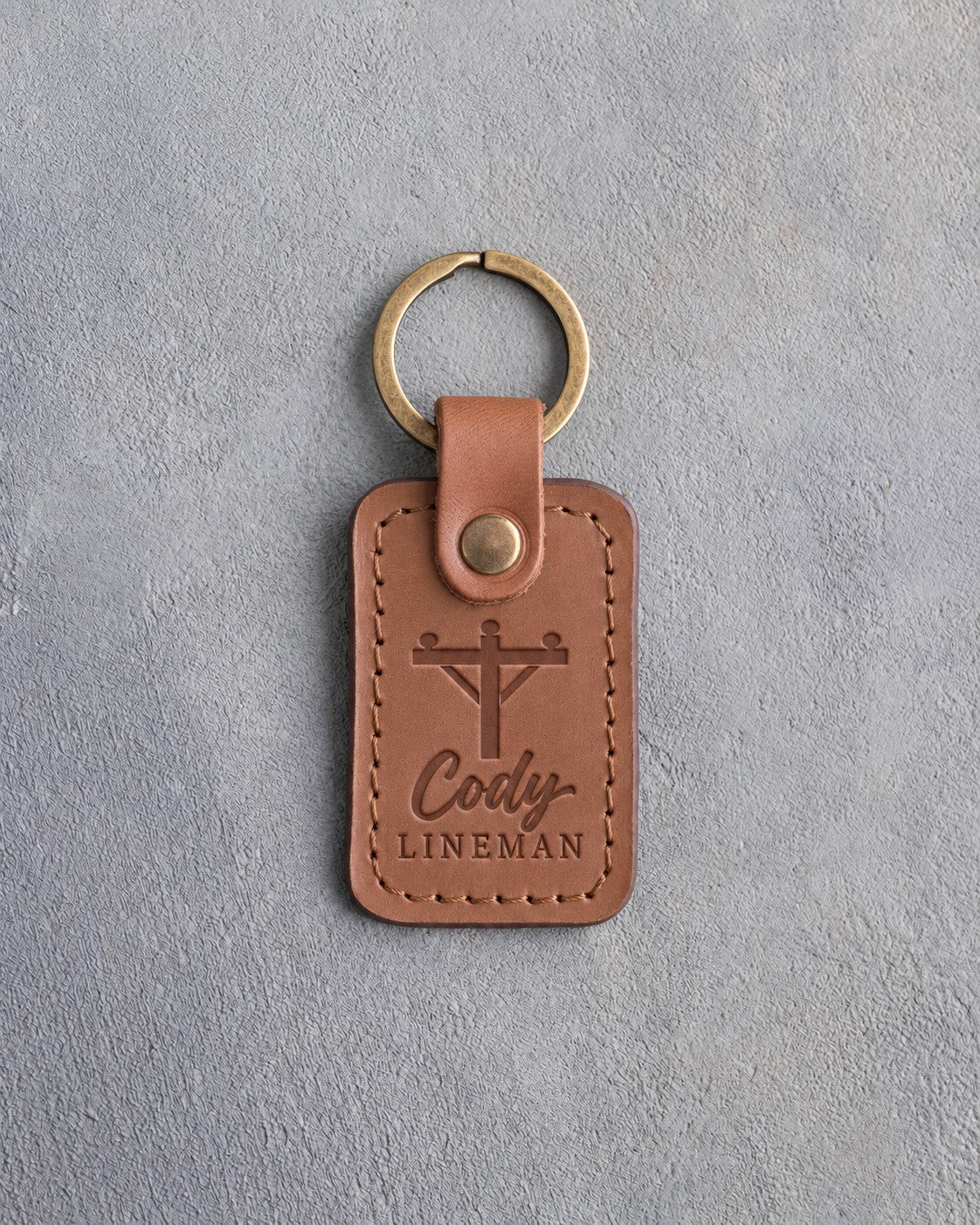 Caramel leather keychain debossed with a utility pole and crossarm above the name Cody and LINEMAN, on gray plaster