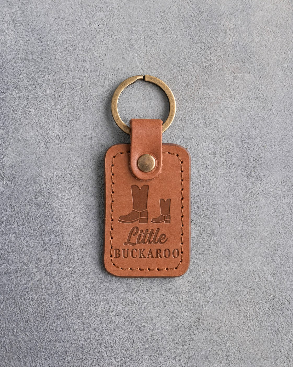 Caramel leather keychain debossed with a big and a small cowboy boot above Little and BUCKAROO, on gray plaster