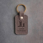 Country gray leather keychain debossed with a big and a small cowboy boot above Little and COWBOY, on gray concrete