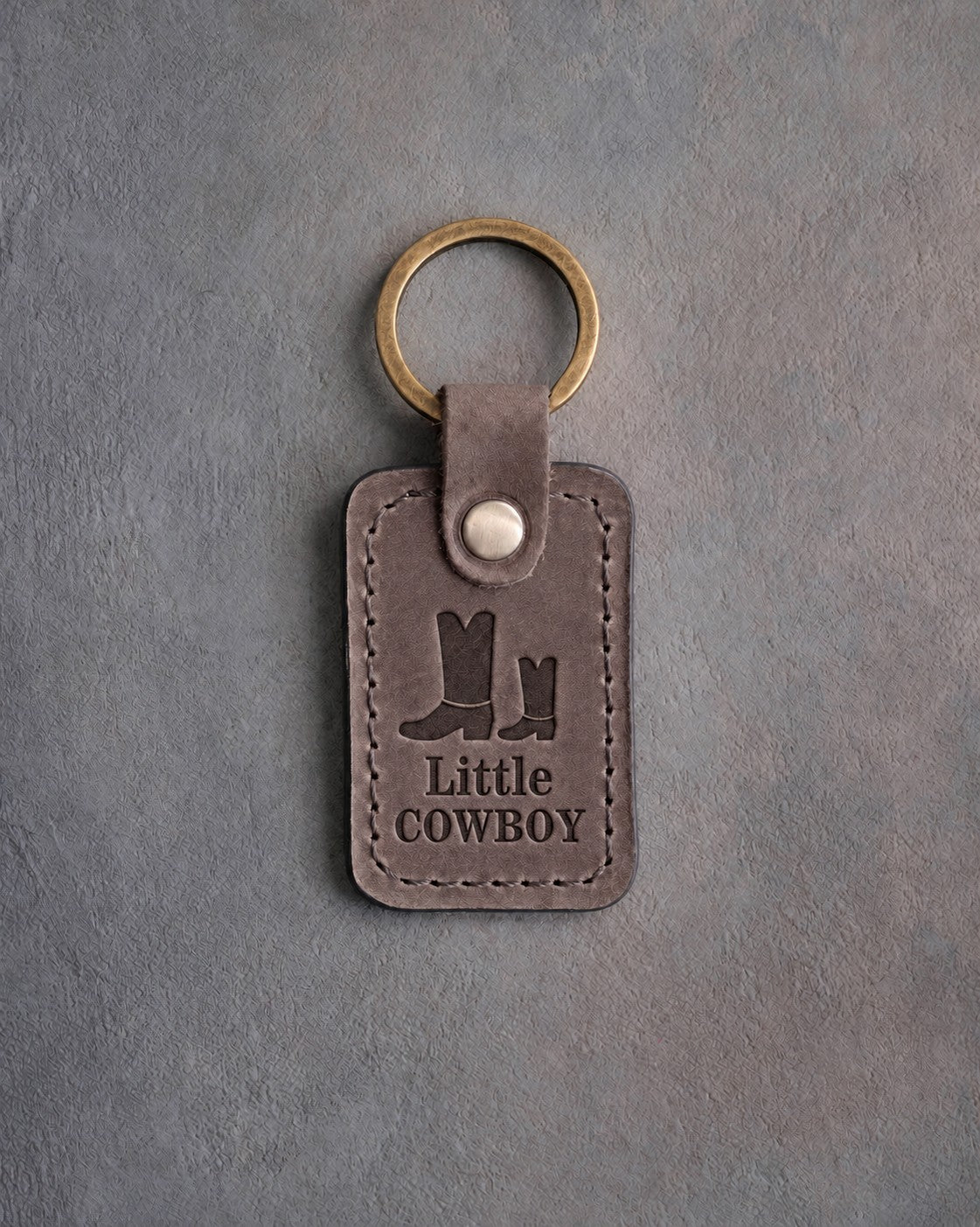 Country gray leather keychain debossed with a big and a small cowboy boot above Little and COWBOY, on gray concrete
