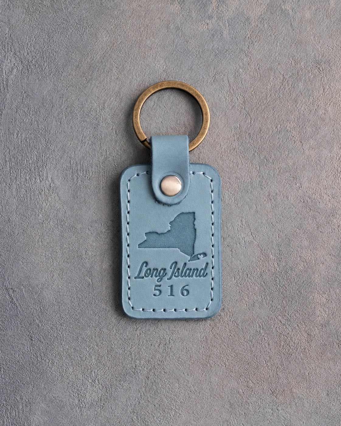 Turquoise leather keychain debossed with a New York State silhouette and a heart notch on Long Island above Long Island and 516, on gray concrete