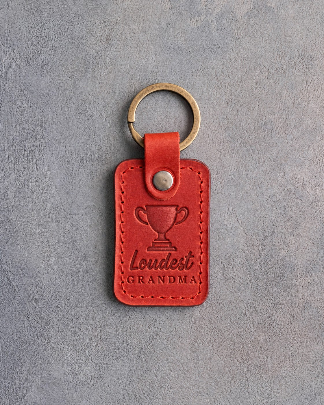 Poppy red leather keychain debossed with a trophy cup and the words Loudest GRANDMA, on gray plaster
