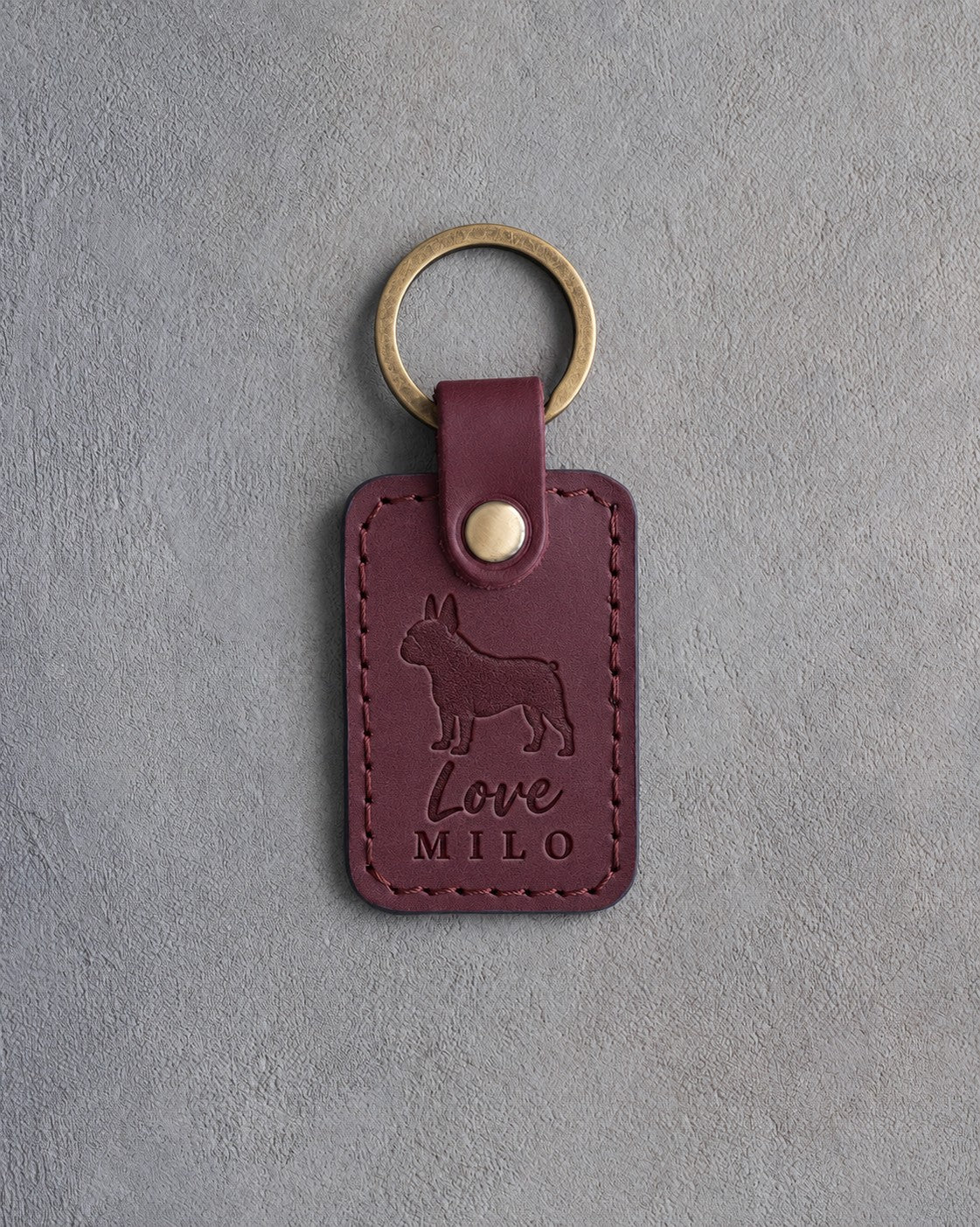 Sangria leather keychain debossed with a French bulldog above the words Love and MILO, on gray concrete