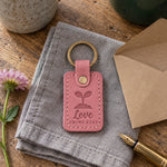 Pink Love Grows Here keychain on a new house key resting on a moving box beside a small potted seedling