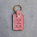 Parisian blossom pink leather keychain debossed with a seedling above Love GROWS HERE, on gray concrete
