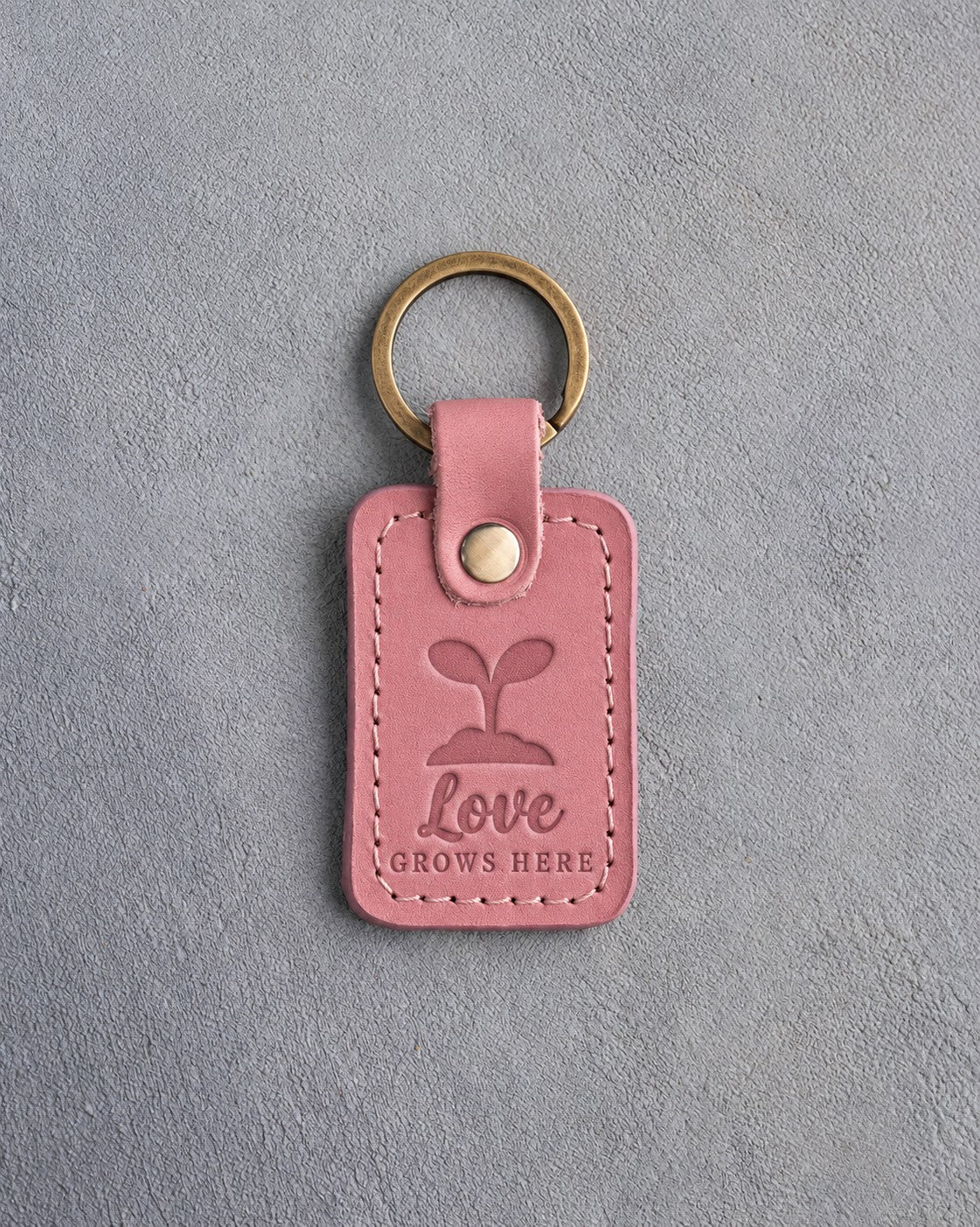 Parisian blossom pink leather keychain debossed with a seedling above Love GROWS HERE, on gray concrete