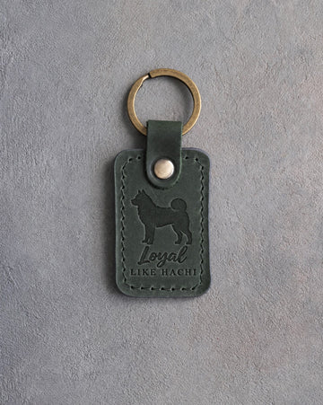 Forest green leather keychain debossed with an Akita and the words Loyal LIKE HACHI, on gray concrete