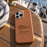 Caramel Grayson MAHI MAHI iPhone case in full-grain leather on the worn teak cockpit table of a sailboat beside a coiled white dock line