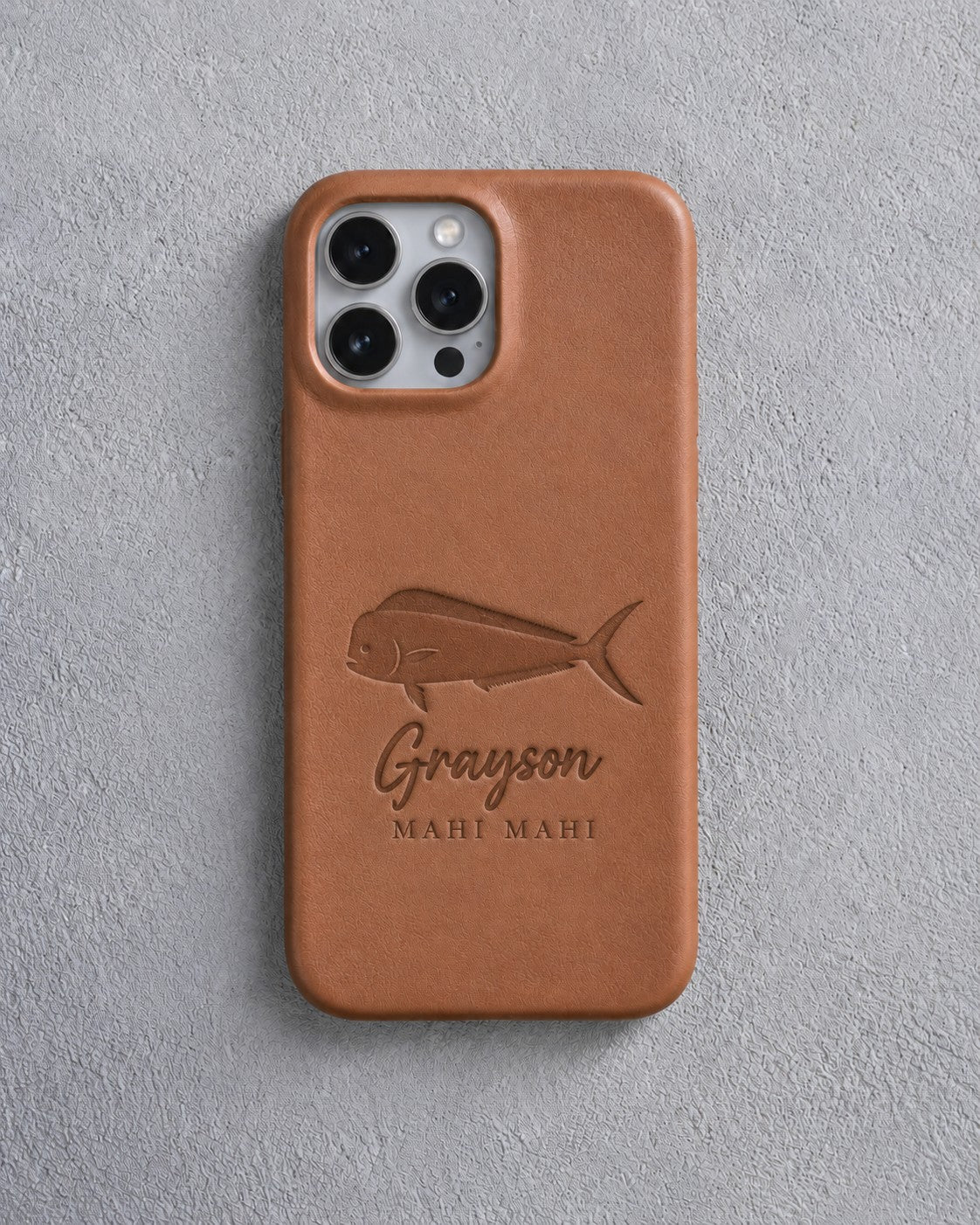 Back of a caramel leather iPhone case debossed with a mahi mahi above Grayson and MAHI MAHI below the camera, on gray plaster