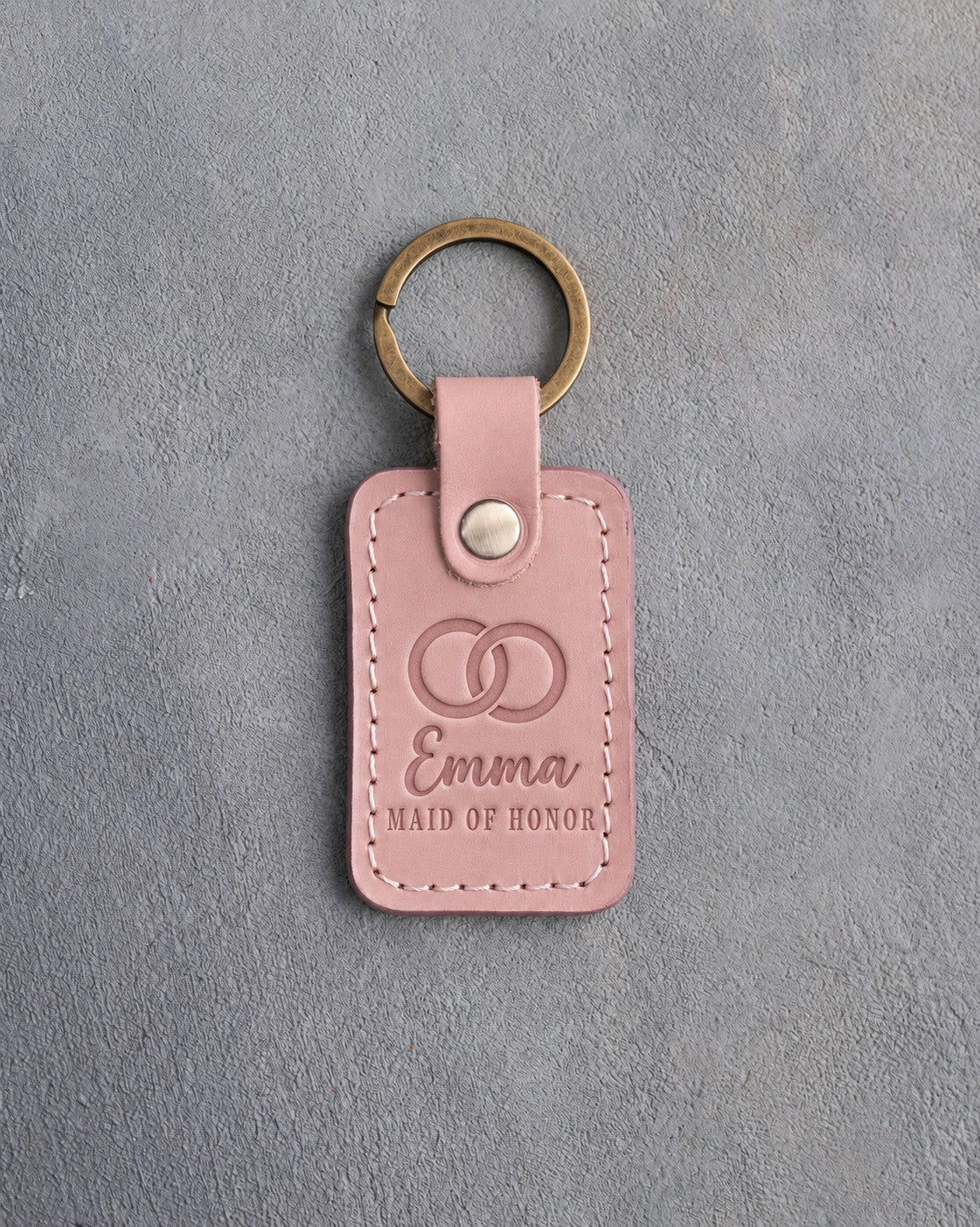 Blush pink leather keychain debossed with two interlocked wedding rings above the name Emma and MAID OF HONOR, on gray concrete