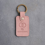 Blush pink leather keychain debossed with two interlocked wedding rings above My and MAID OF HONOR, on gray concrete