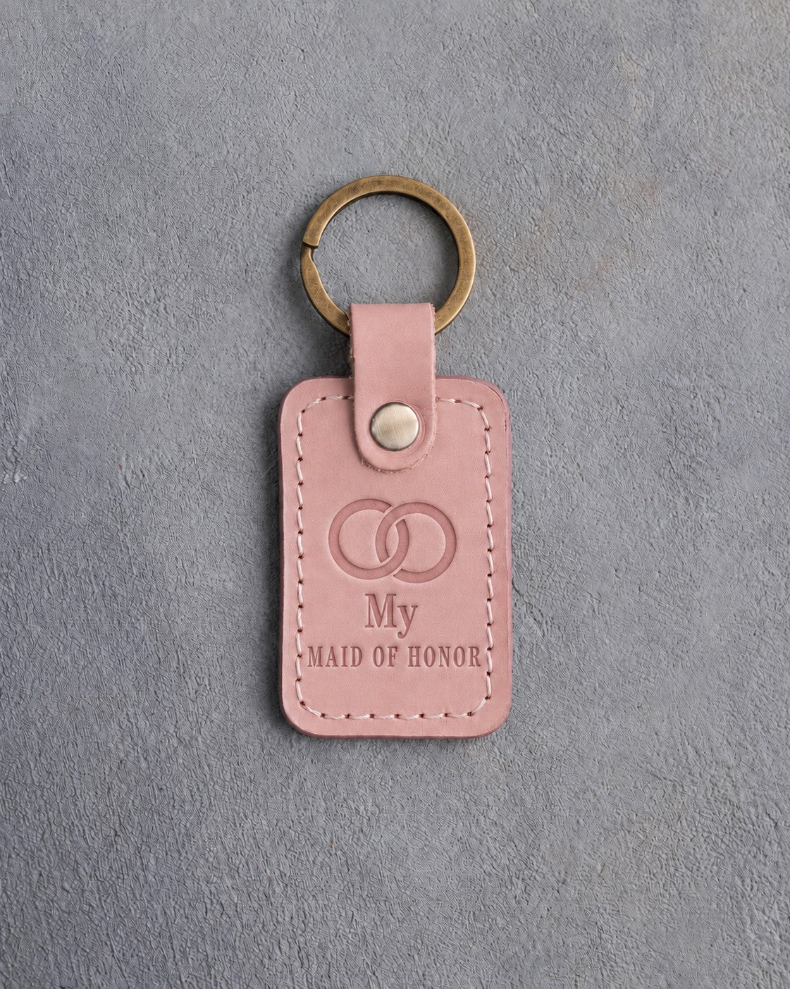 Blush pink leather keychain debossed with two interlocked wedding rings above My and MAID OF HONOR, on gray concrete