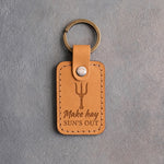 Indian Summer tan leather keychain debossed with an upright pitchfork above the words Make hay SUN'S OUT, on gray concrete