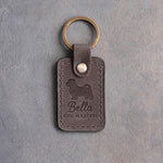 Country gray leather keychain debossed with a Maltese silhouette, the name Bella and THE MALTESE, on gray concrete