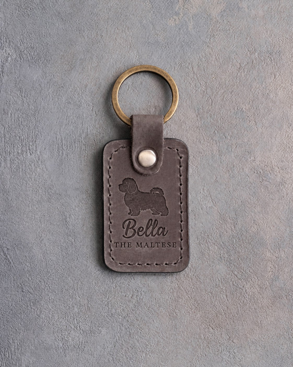 Country gray leather keychain debossed with a Maltese silhouette, the name Bella and THE MALTESE, on gray concrete