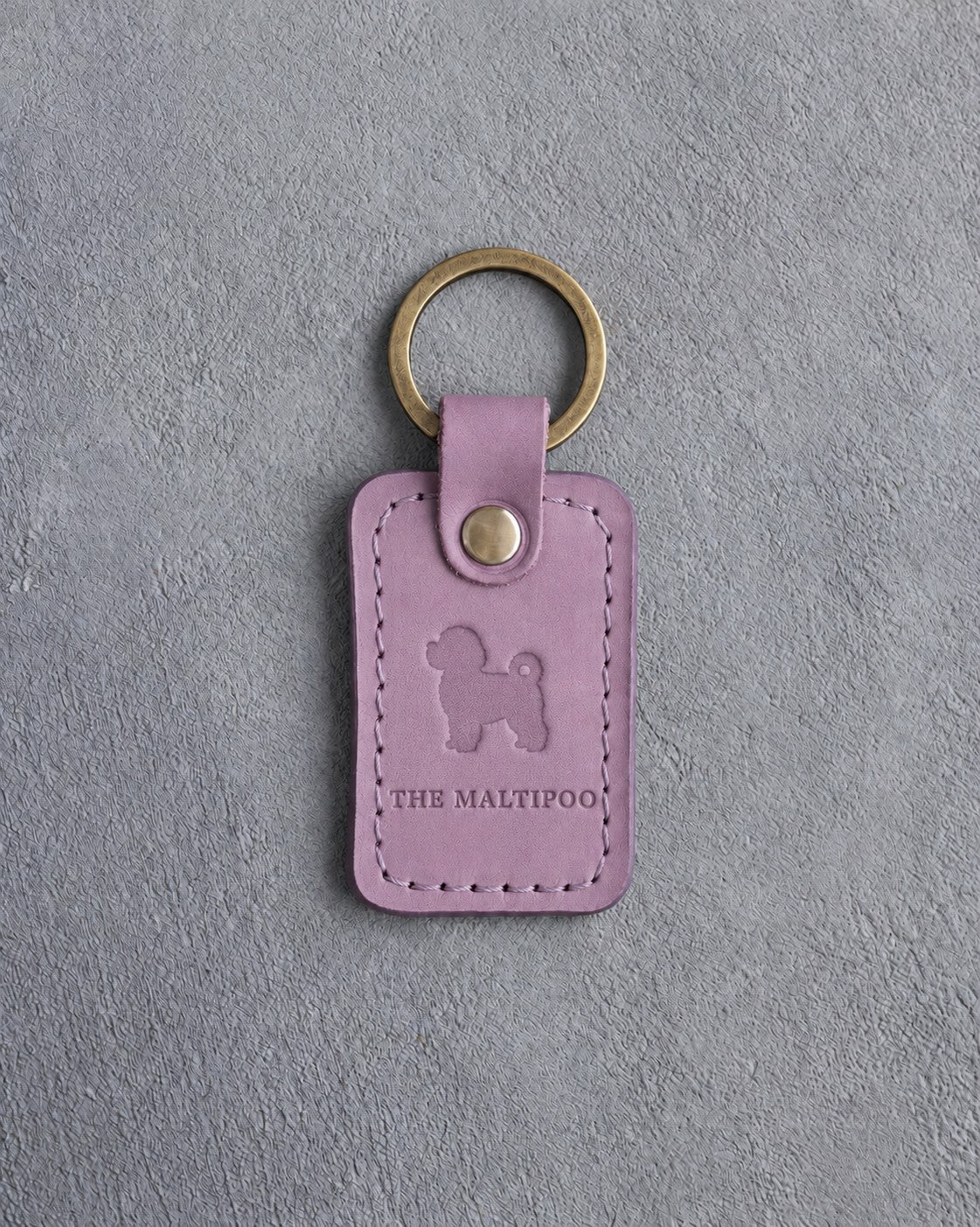 Lilac leather keychain debossed with a tiny maltipoo above THE MALTIPOO, on gray concrete