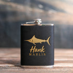 Black leather-wrapped hip flask stamped in gold foil with a blue marlin above Hank and MARLIN, on a wooden table