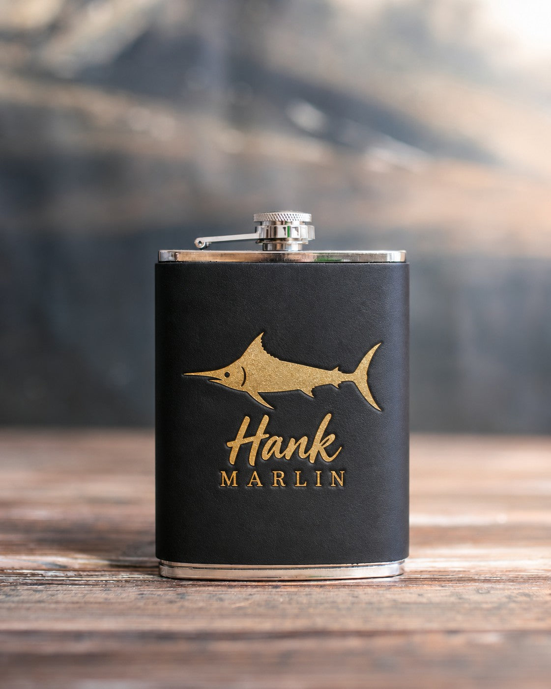 Black leather-wrapped hip flask stamped in gold foil with a blue marlin above Hank and MARLIN, on a wooden table