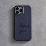 Back of a blueberry leather iPhone case debossed with a blue marlin above Hank and MARLIN below the camera, on gray plaster