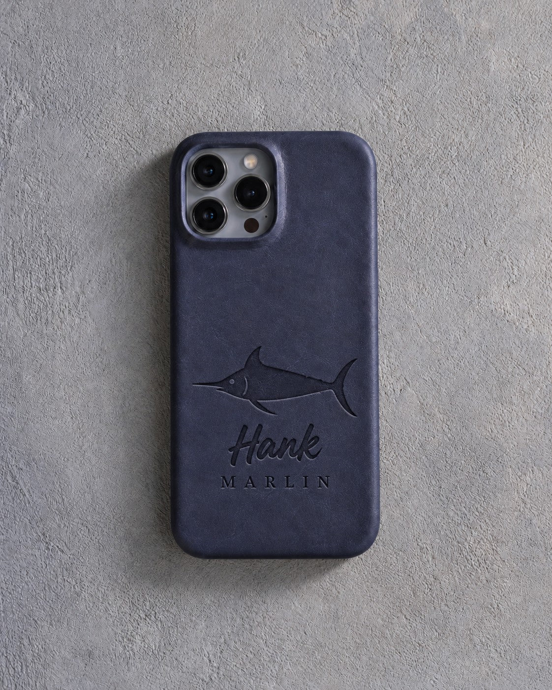 Back of a blueberry leather iPhone case debossed with a blue marlin above Hank and MARLIN below the camera, on gray plaster