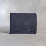 Closed blueberry leather bifold wallet with a blue marlin beside Hank and MARLIN pressed in the lower right corner, on gray plaster