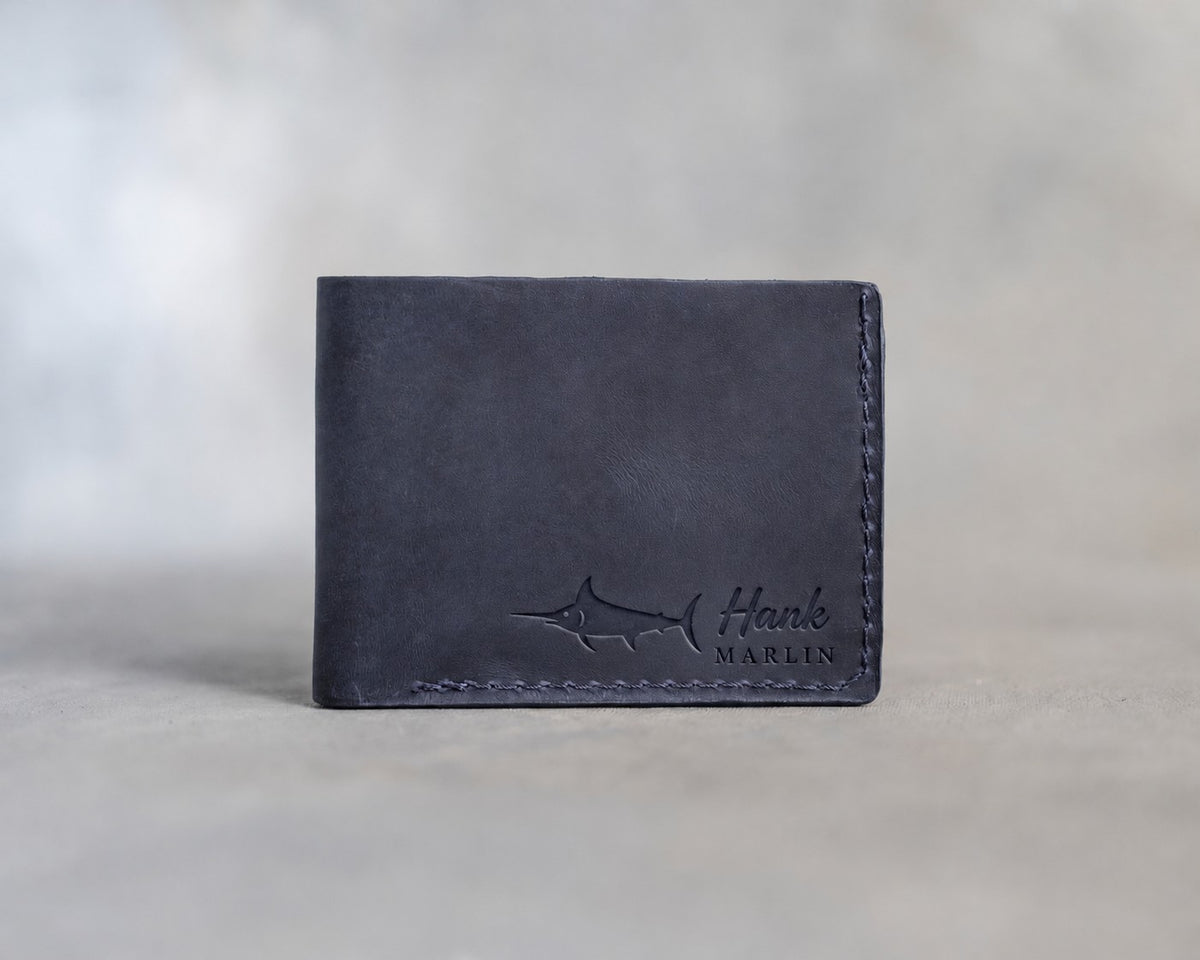 Closed blueberry leather bifold wallet with a blue marlin beside Hank and MARLIN pressed in the lower right corner, on gray plaster