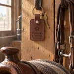 Sicilian brown Married A COWGIRL keychain hanging from an iron bridle hook in a tack room, a saddle soft on its rack below