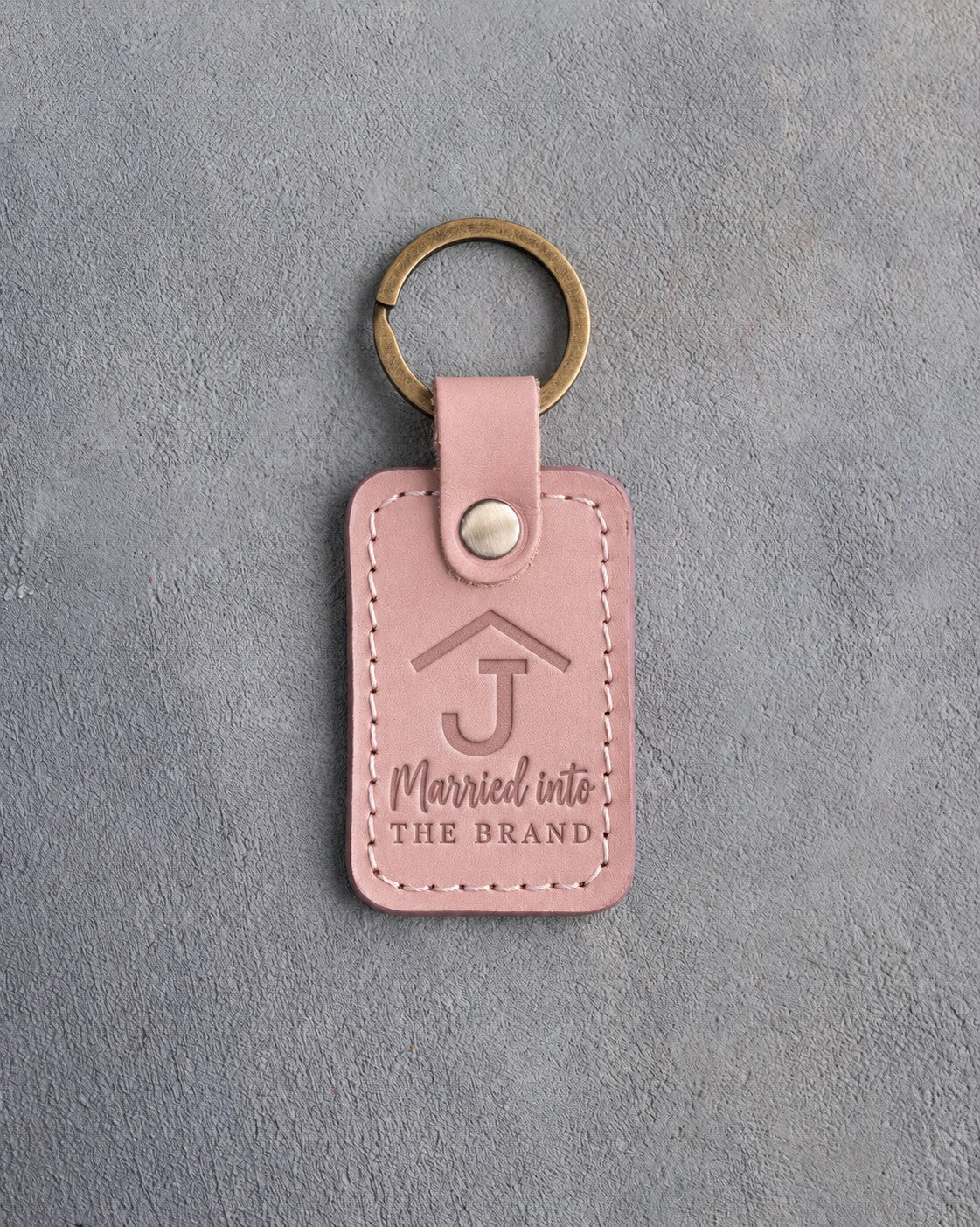 Blush pink leather keychain debossed with a Rafter J ranch brand above Married into and THE BRAND, on gray plaster
