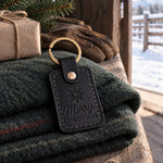 Black Merry CHRISTMAS keychain with a gold rowel on a folded green wool blanket inside a barn door, snow on the corral rails outside