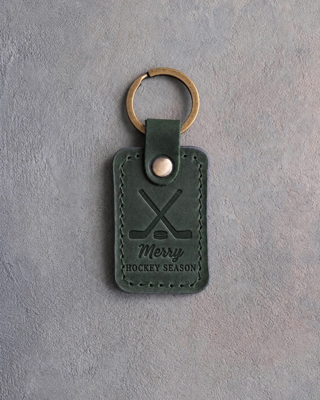 Forest green leather keychain debossed with crossed hockey sticks and a puck and the words Merry HOCKEY SEASON, on gray plaster