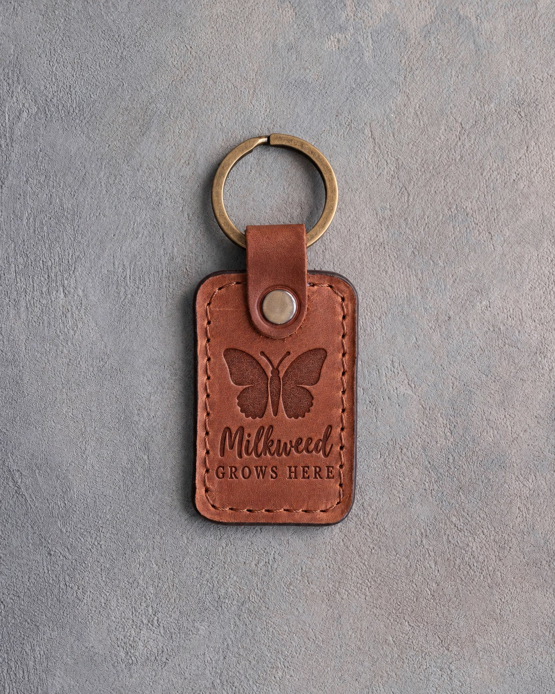 Arizona tan leather keychain debossed with a butterfly above Milkweed GROWS HERE, on gray concrete