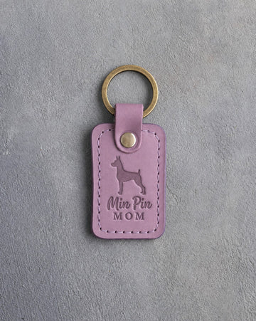 Lilac leather keychain debossed with a miniature pinscher and the words Min Pin MOM, on gray concrete