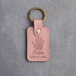 Blush pink leather keychain debossed with a small hand inside a large hand above Mom and HAND IN HAND, on gray concrete