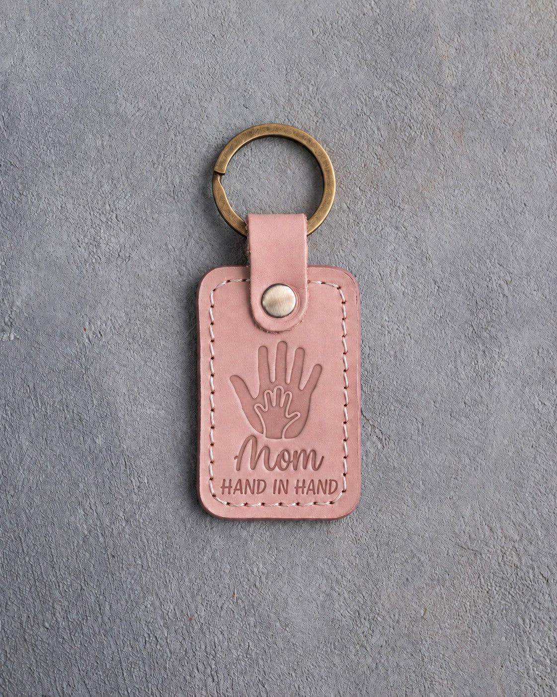 Blush pink leather keychain debossed with a small hand inside a large hand above Mom and HAND IN HAND, on gray concrete