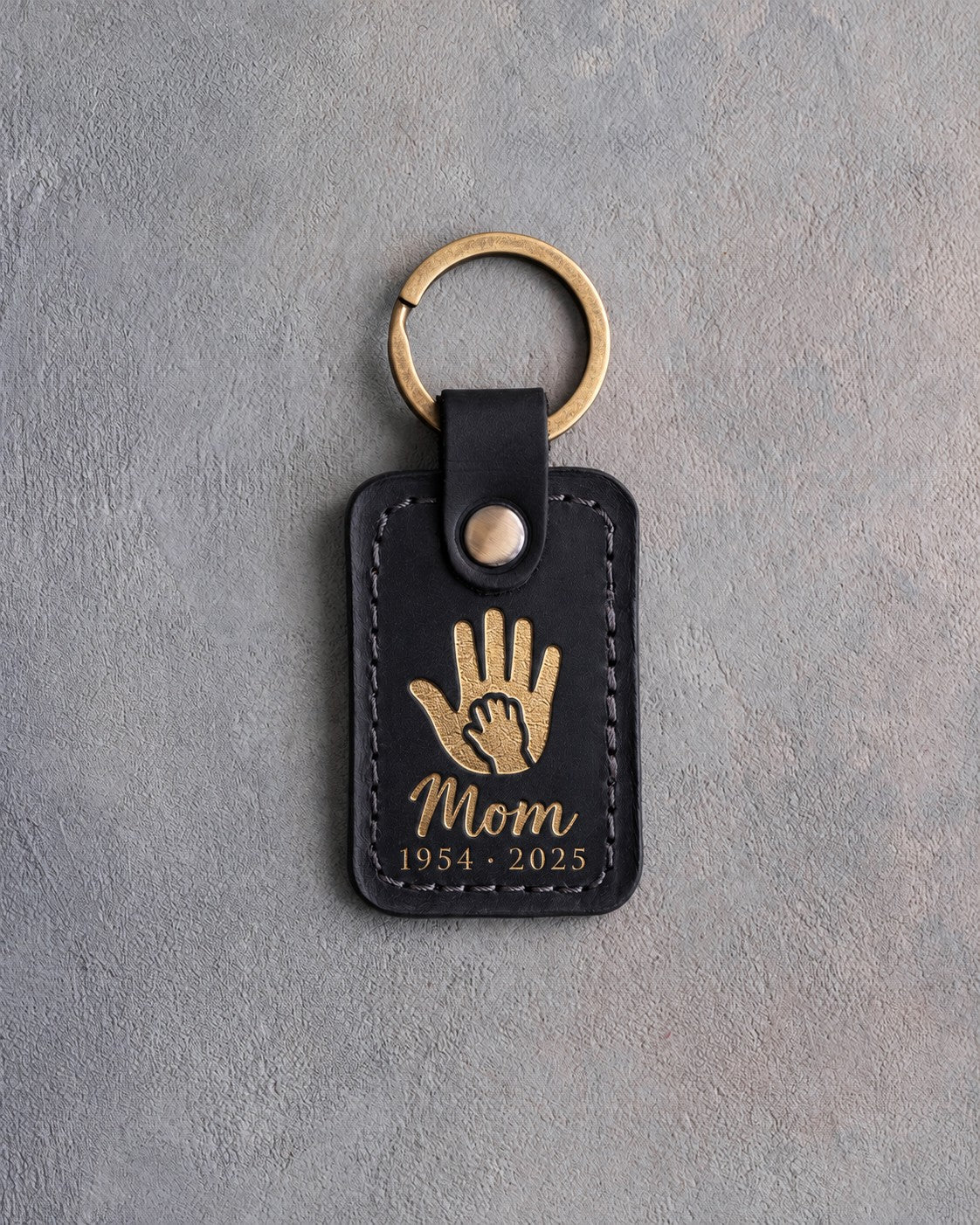 Black leather keychain with a gold foil small hand inside a large hand above Mom and 1954 · 2025