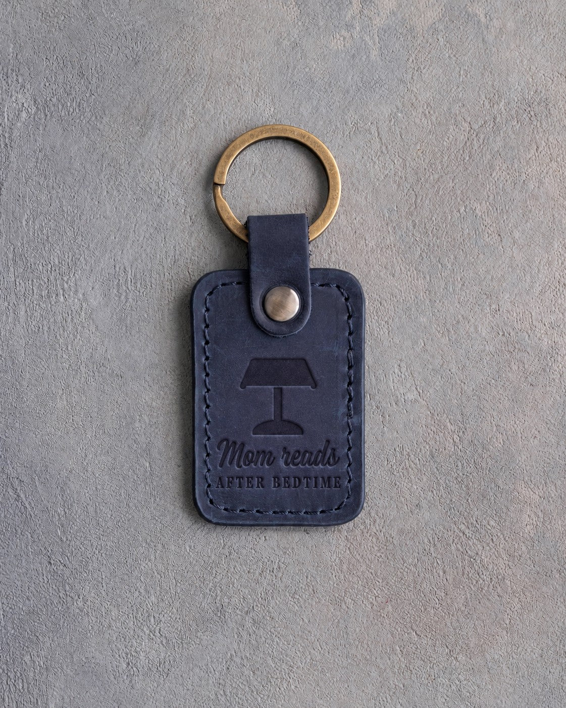 Blueberry navy leather keychain debossed with a banker's lamp above Mom reads and AFTER BEDTIME, on gray concrete