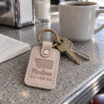 Pale gray Montana Raised Me keychain on truck keys resting on a fence post with open prairie behind