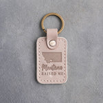 Gray sand leather keychain debossed with a wide Montana silhouette and a heart at Bozeman above Montana and RAISED ME, on gray concrete