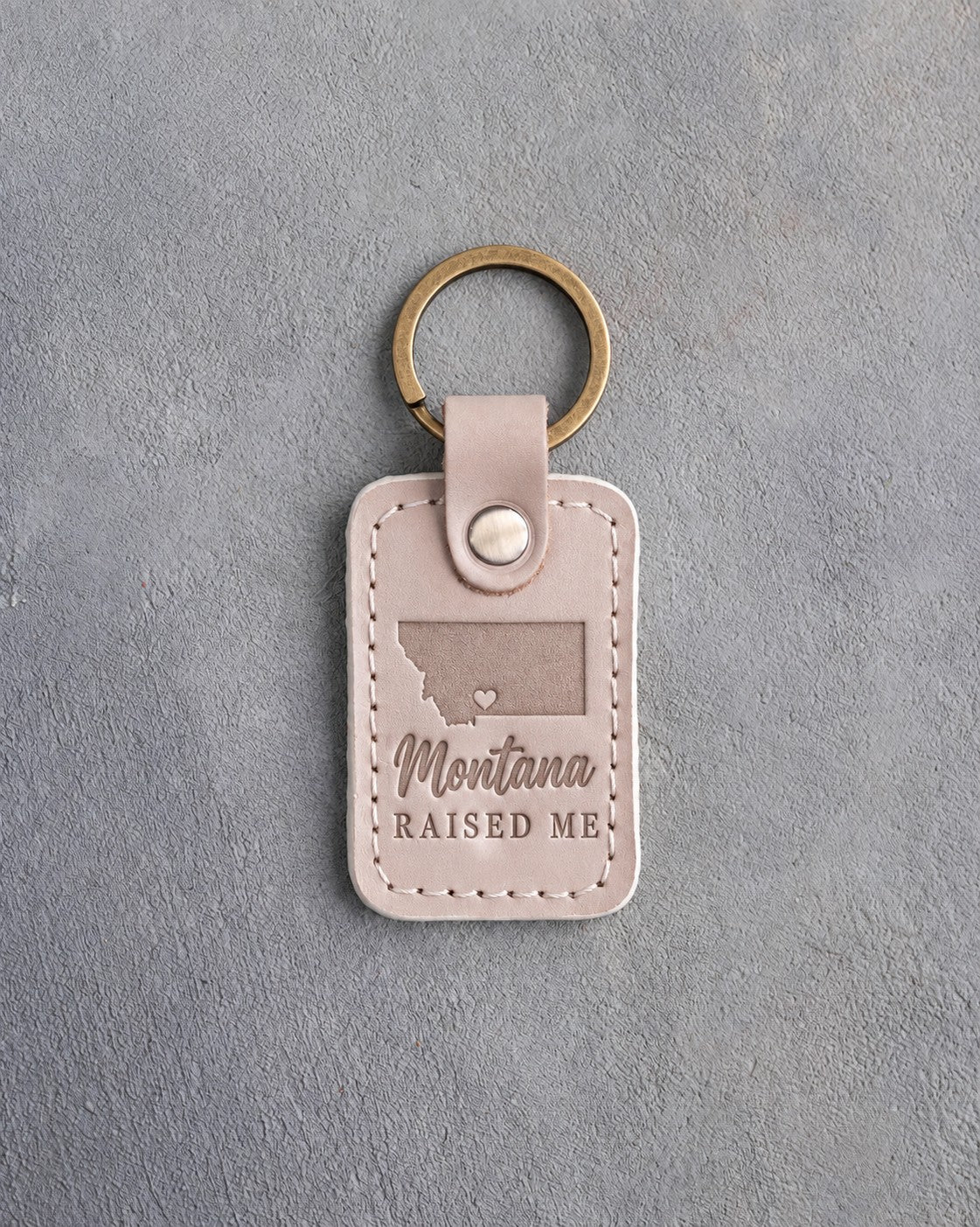 Gray sand leather keychain debossed with a wide Montana silhouette and a heart at Bozeman above Montana and RAISED ME, on gray concrete