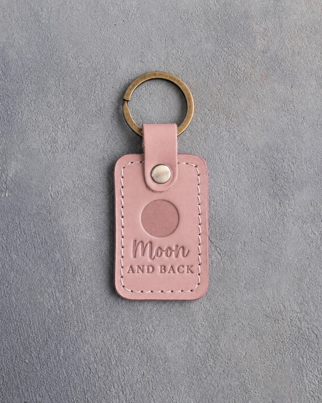 Blush pink leather keychain debossed with a solid full moon above the words Moon AND BACK, on gray concrete