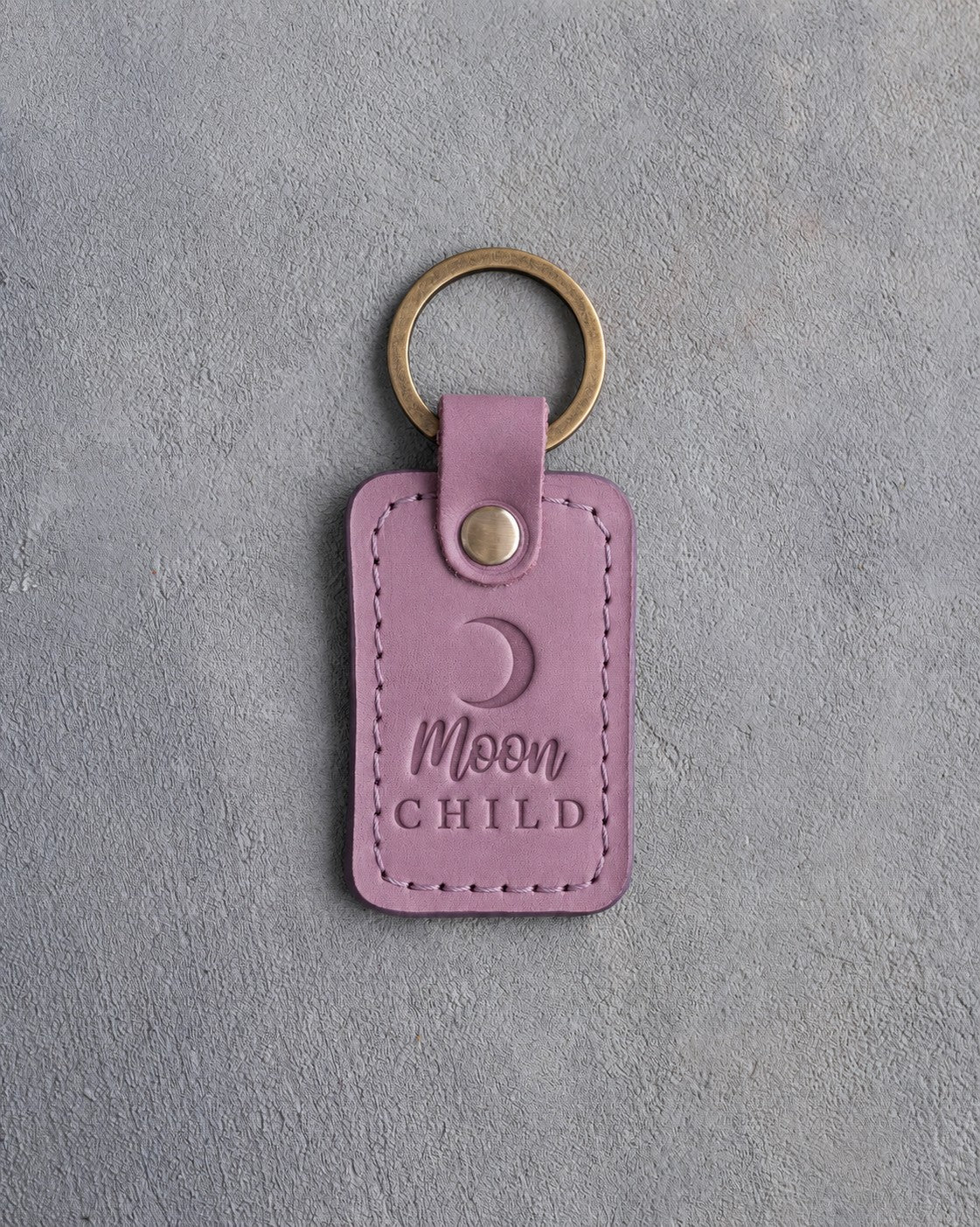 Lilac leather keychain debossed with a waxing crescent moon above the words Moon CHILD, on gray concrete